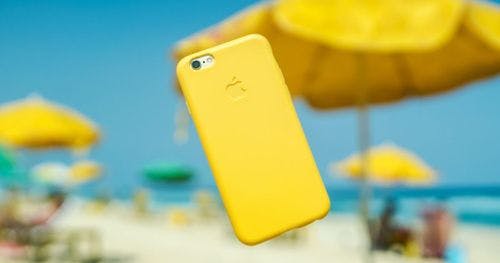 Smartphone mit gelber Hülle im Vordergrund, vor einem Strand mit gelbem Sonnenschirm